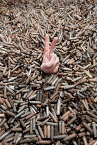 Hand making a victory gesture with two fingers in the foreground, while bullet shells are scattered in the background, representing war and the desire for peace."